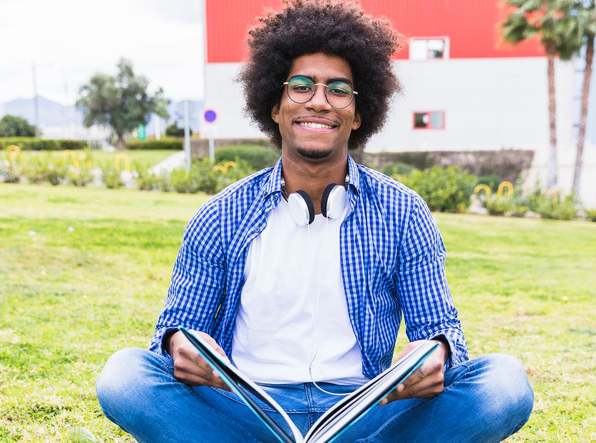 A happy student sitting on the ground inside a university campus, engaged in studies.